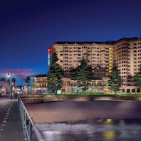 Hotel Stamford Grand Adelaide Glenelg Exterior foto The photo shows a waterfront scene at dusk, featuring a large building lit up against a darkening sky. The architecture appears modern and prominent, with several windows illuminated. In the foreground, there is a walkway or pier with fencing along t