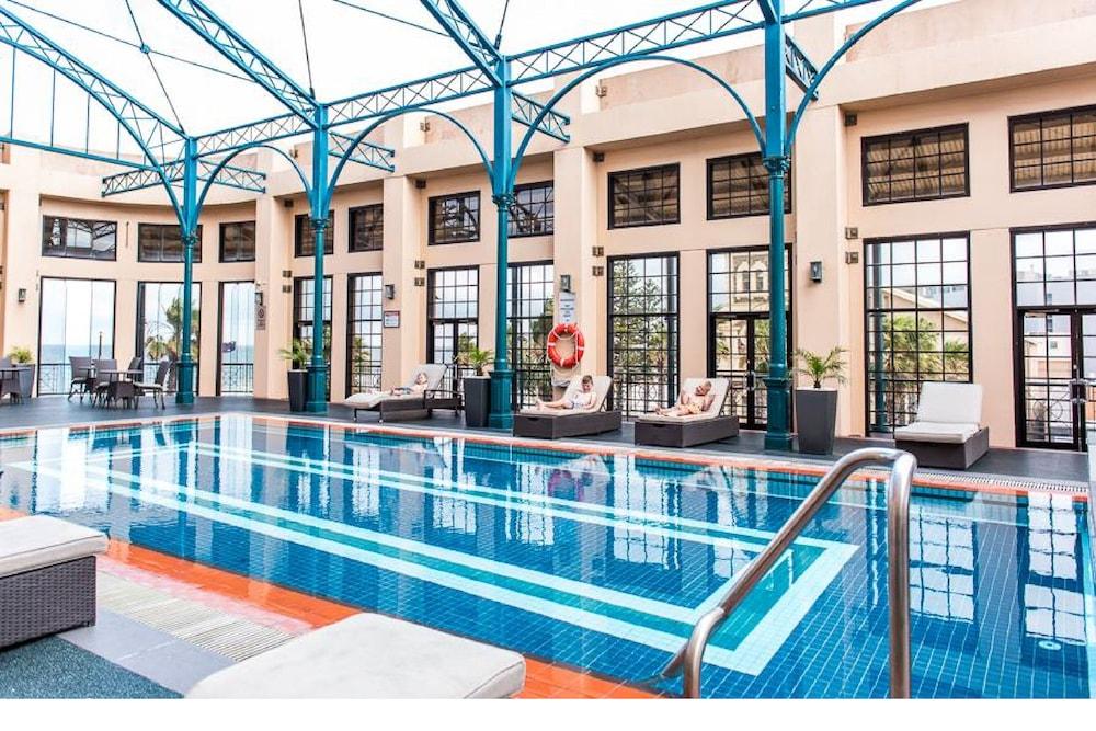 Hotel Stamford Grand Adelaide Glenelg Exterior foto The photo shows an indoor swimming pool area characterized by a modern and elegant design. The pool features blue tiles with two white stripes running along its length. Surrounding the pool, there are several lounge chairs for relaxation. The walls h