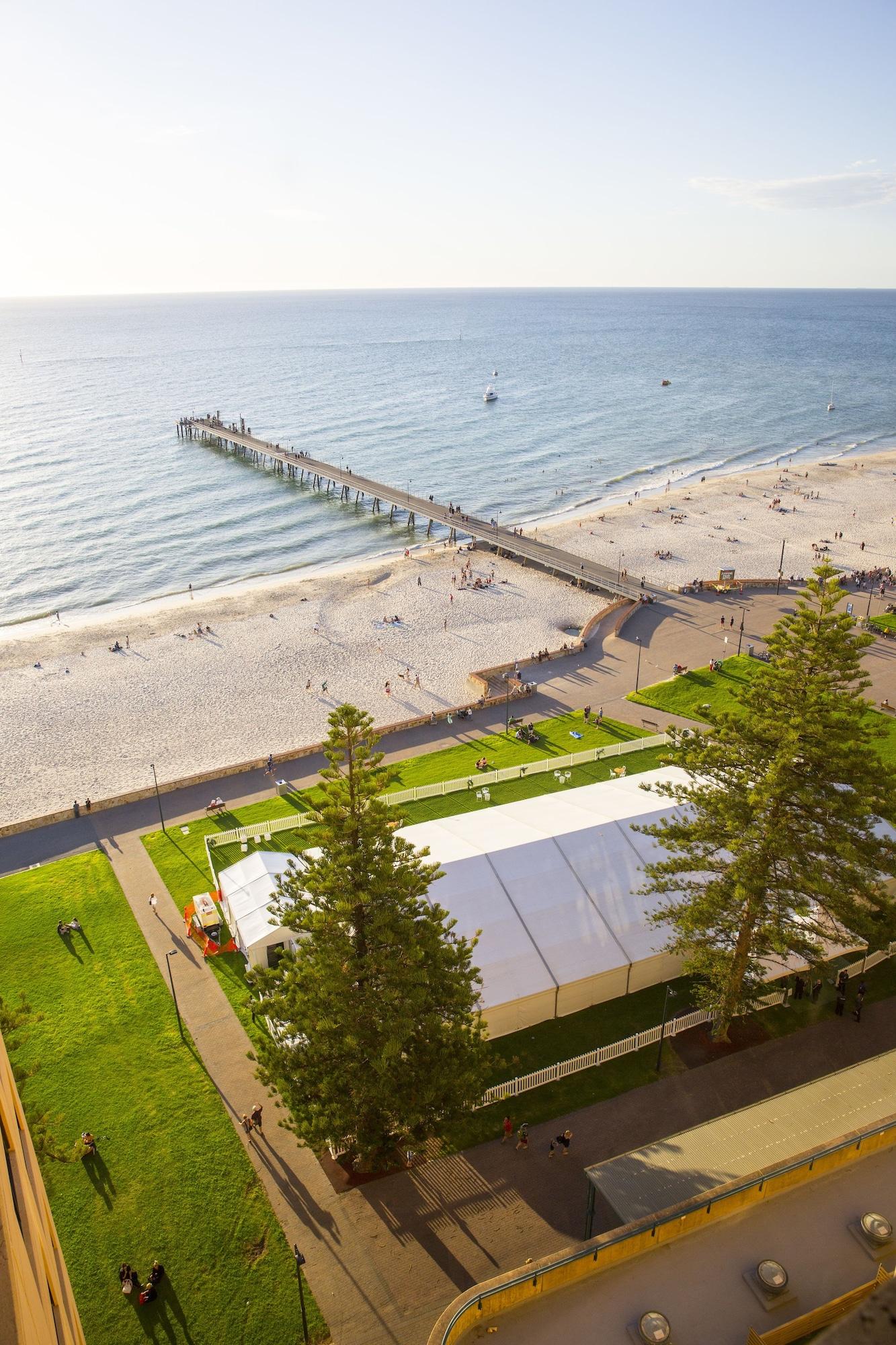 Hotel Stamford Grand Adelaide Glenelg Exterior foto The photo shows a scenic view of a beach with a long pier extending into the water. The beach is sandy, and there are people enjoying the sun and walking along the shore. In the foreground, there is a large white tent set up on a grassy area, likely 