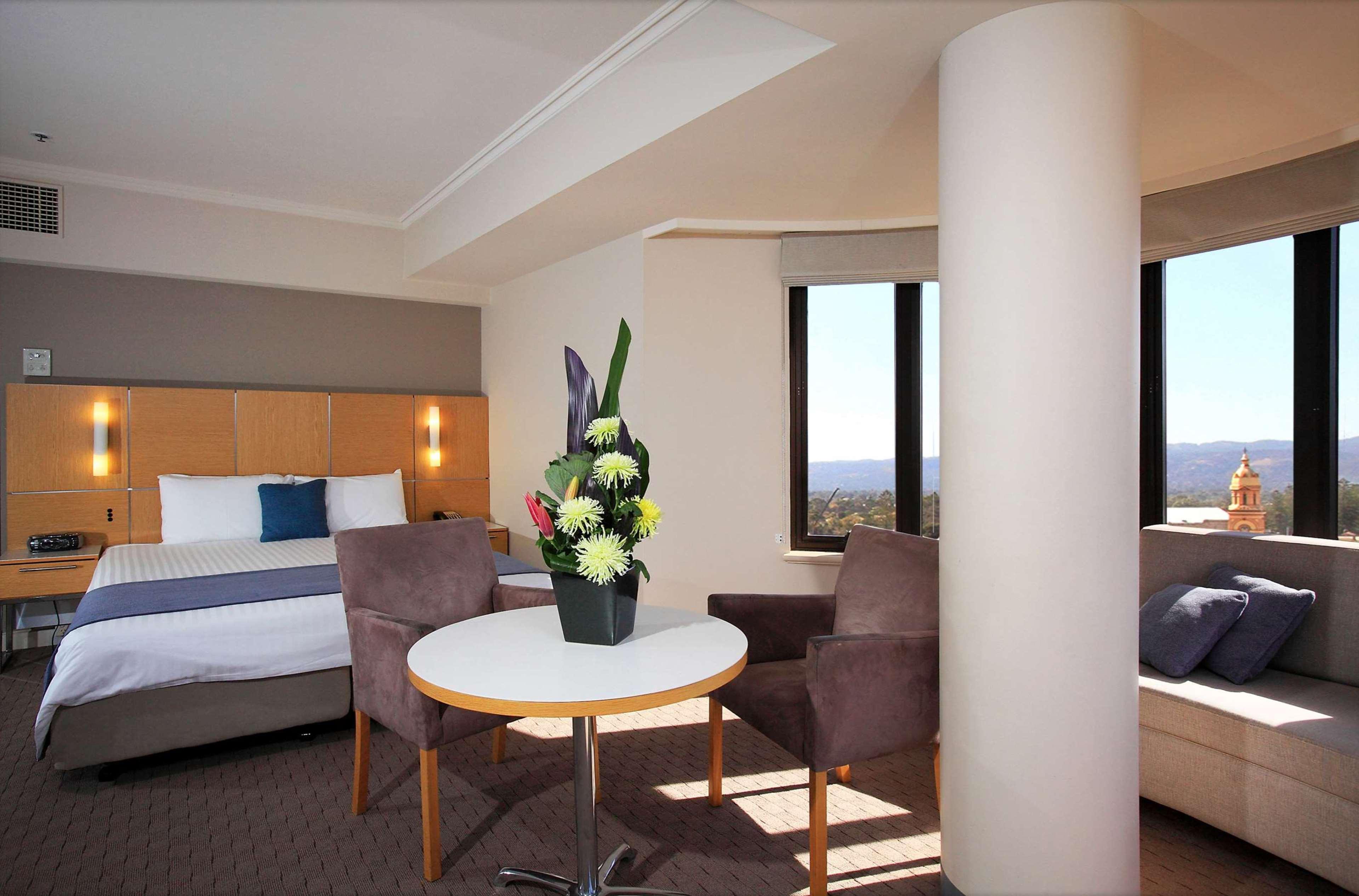 Hotel Stamford Grand Adelaide Glenelg Exterior foto The photo shows a hotel room with a modern design. On one side, there is a plush bed with white and blue bedding. In the center of the room, there's a small round table with two gray upholstered chairs around it. A vase of flowers sits on the table, 