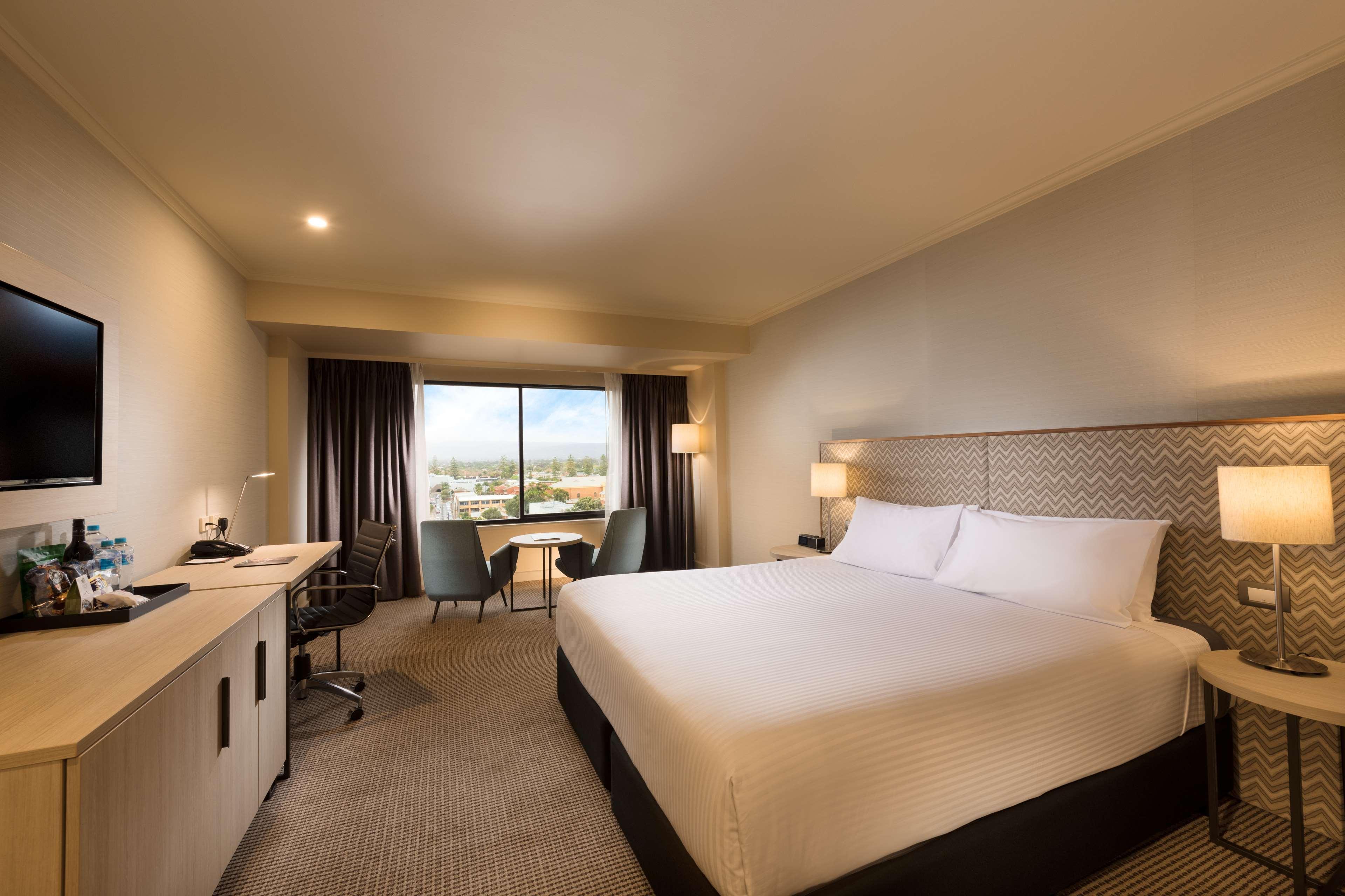 Hotel Stamford Grand Adelaide Glenelg Exterior foto The photo shows a modern hotel room. It features a large bed with white linens, a desk with a chair, and a flat-screen TV mounted on the wall. There are two armchairs and a small table near a large window that lets in natural light, providing a view 