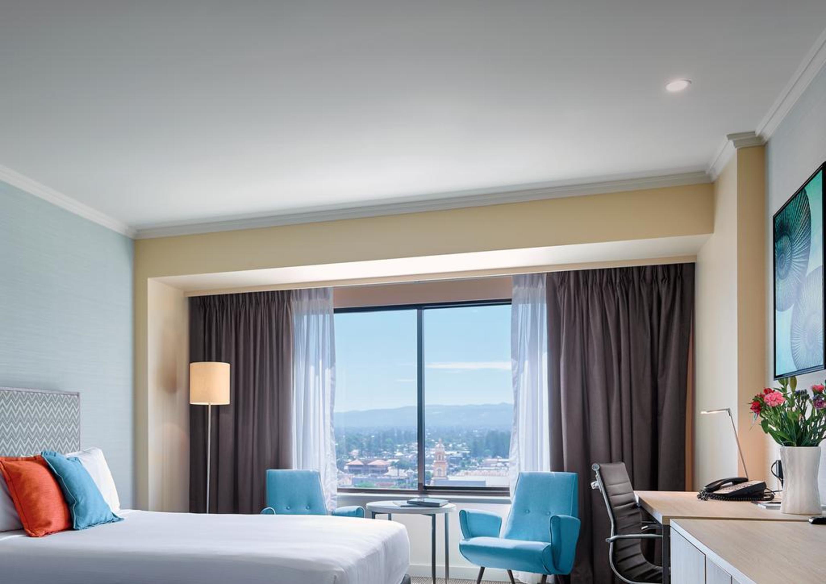 Hotel Stamford Grand Adelaide Glenelg Exterior foto The photo shows a modern hotel room featuring a queen-sized bed with white linens and colorful decorative pillows. There are two blue chairs beside a small table positioned near a large window that provides a view of the outside, with sheer and dark 