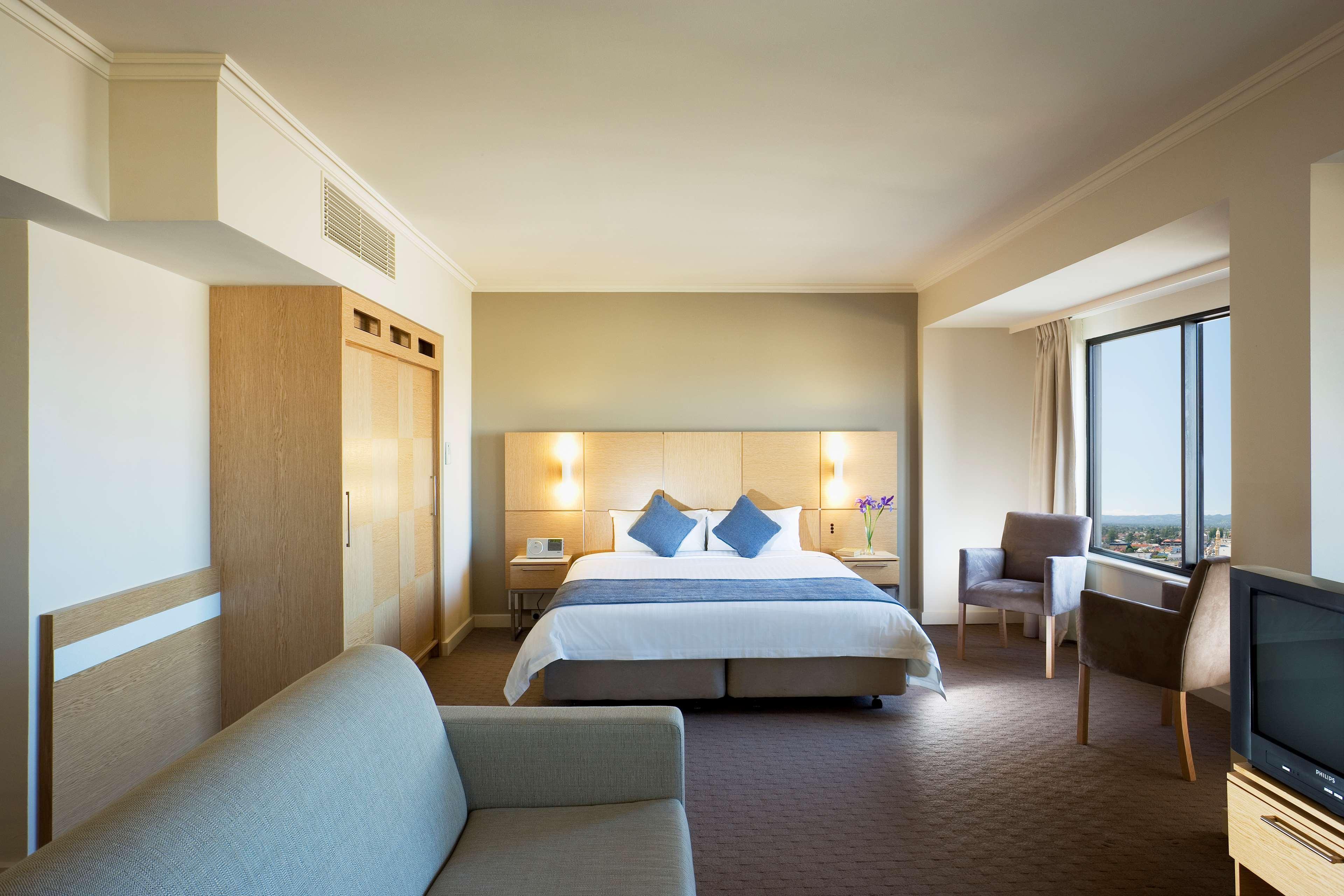 Hotel Stamford Grand Adelaide Glenelg Zimmer foto The photo shows a modern hotel room featuring a large bed with two blue decorative pillows. There are bedside lamps on both sides of the headboard. To the left, there's a wooden wardrobe and a small table with a vase and a few amenities. On the right