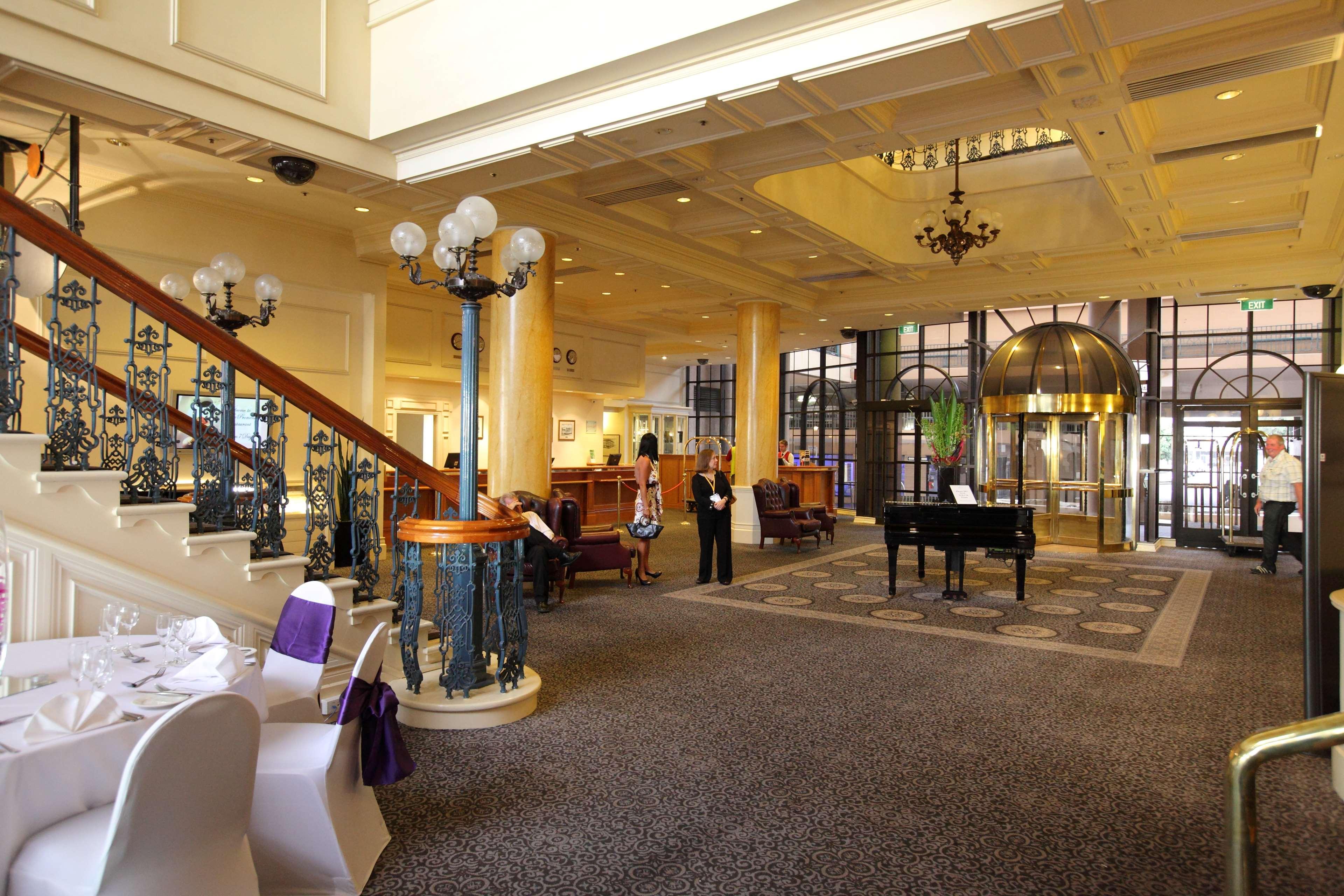 Hotel Stamford Grand Adelaide Glenelg Interior foto The photo shows a luxurious interior of a hotel or a restaurant lobby. There is an elegant staircase with ornate railings leading to an upper level. The space features high ceilings with decorative chandeliers and ample natural light coming through l