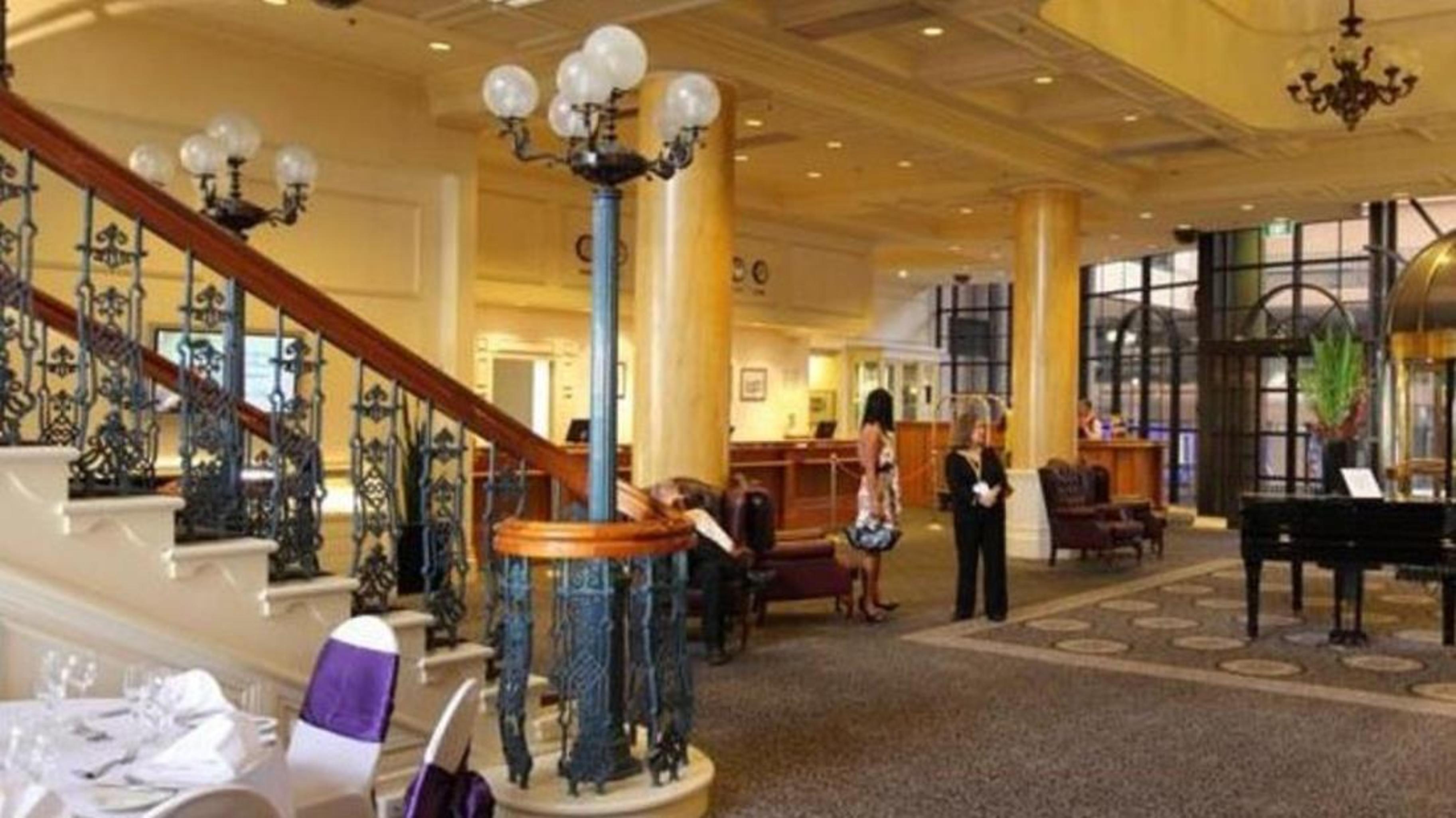 Hotel Stamford Grand Adelaide Glenelg Exterior foto The photo shows the interior of a hotel lobby. It features a spacious area with a high ceiling and elegant decor. There is a staircase with a decorative railing leading to an upper level. In the foreground, some guests and staff are present, and ther