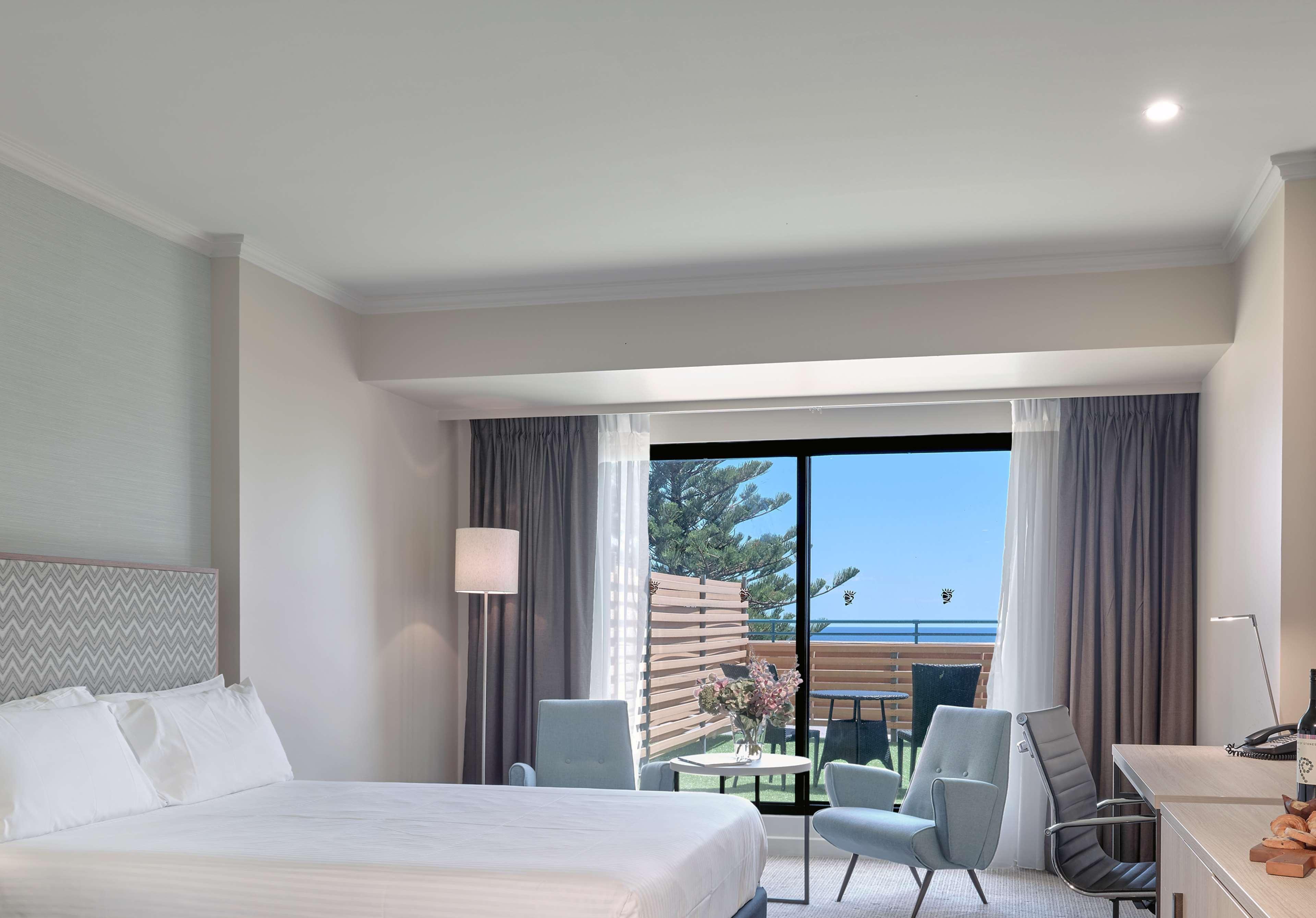 Hotel Stamford Grand Adelaide Glenelg Exterior foto The photo shows a modern hotel room featuring a large bed with white bedding. There are two armchairs positioned near a window that offers a view of an outdoor area, possibly a balcony or patio with greenery. The room has light-colored walls and curt