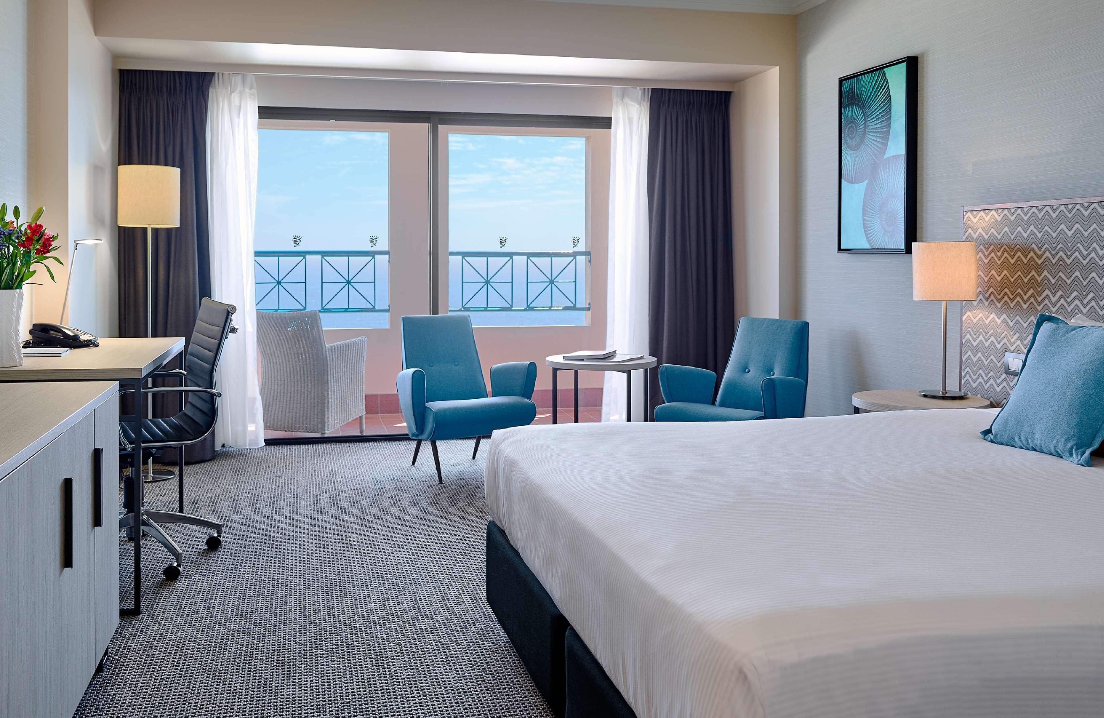 Hotel Stamford Grand Adelaide Glenelg Exterior foto The image shows a hotel room featuring a large bed with a white duvet. There are two bright blue chairs and a small table near a large window that provides a view outside, possibly to a balcony. The room includes a workspace with a desk and an office