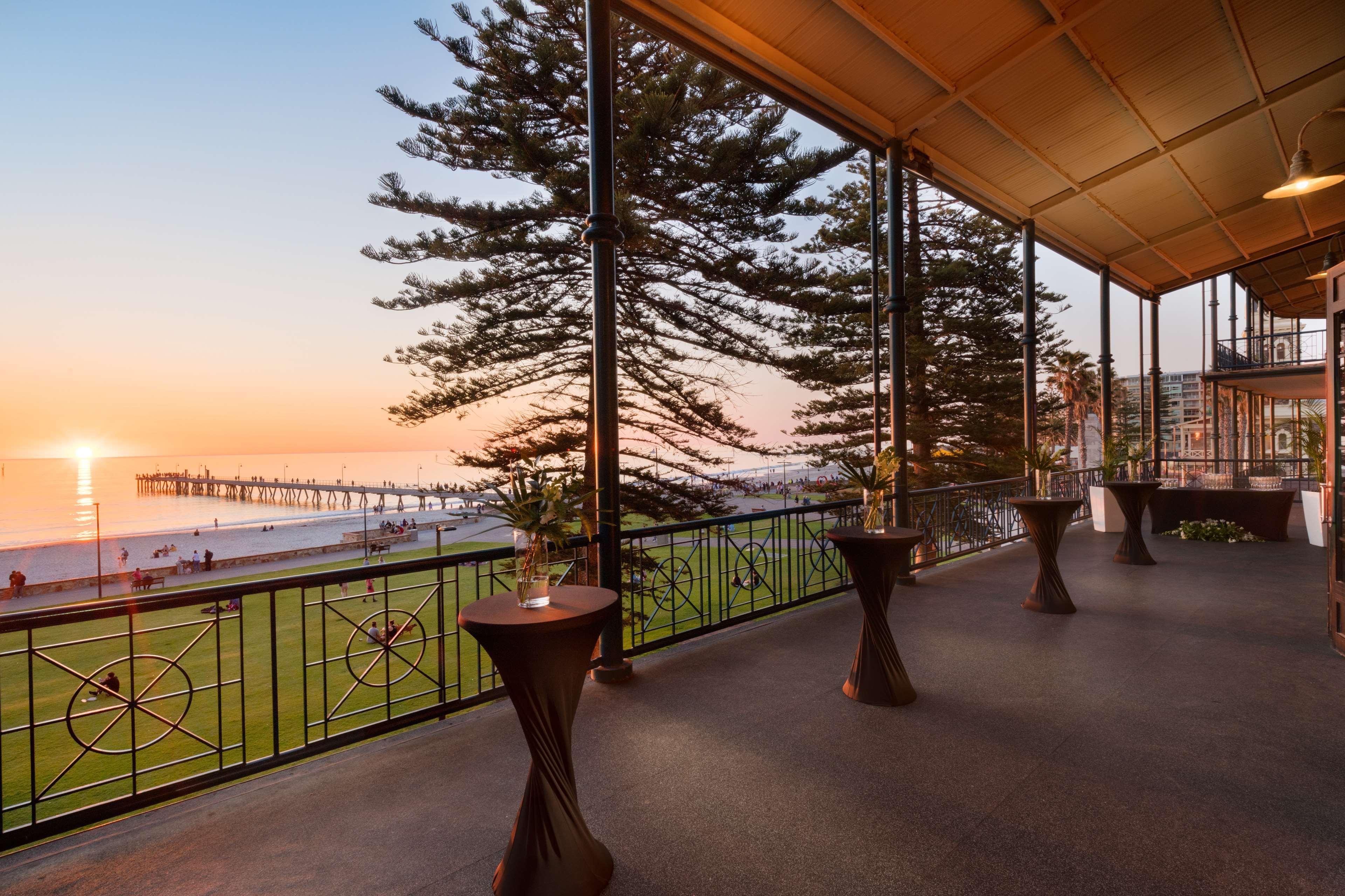 Hotel Stamford Grand Adelaide Glenelg Exterior foto The photo shows a spacious indoor space with large windows overlooking a beach at sunset. The scene features a pier extending into the ocean and a horizon painted in warm colors from the setting sun. In the foreground, there are elegant tables with d