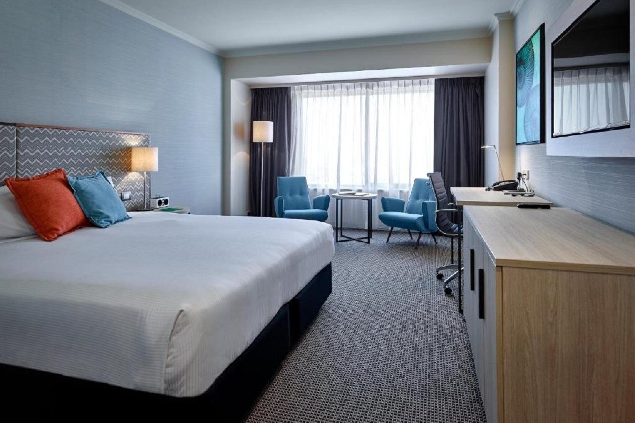 Hotel Stamford Grand Adelaide Glenelg Exterior foto The photo shows a modern hotel room with a clean and minimalist design. In the center, there is a large, neatly made bed with white linens and two decorative pillows, one orange and one blue. To the right, there is a small wooden cabinet with a telev